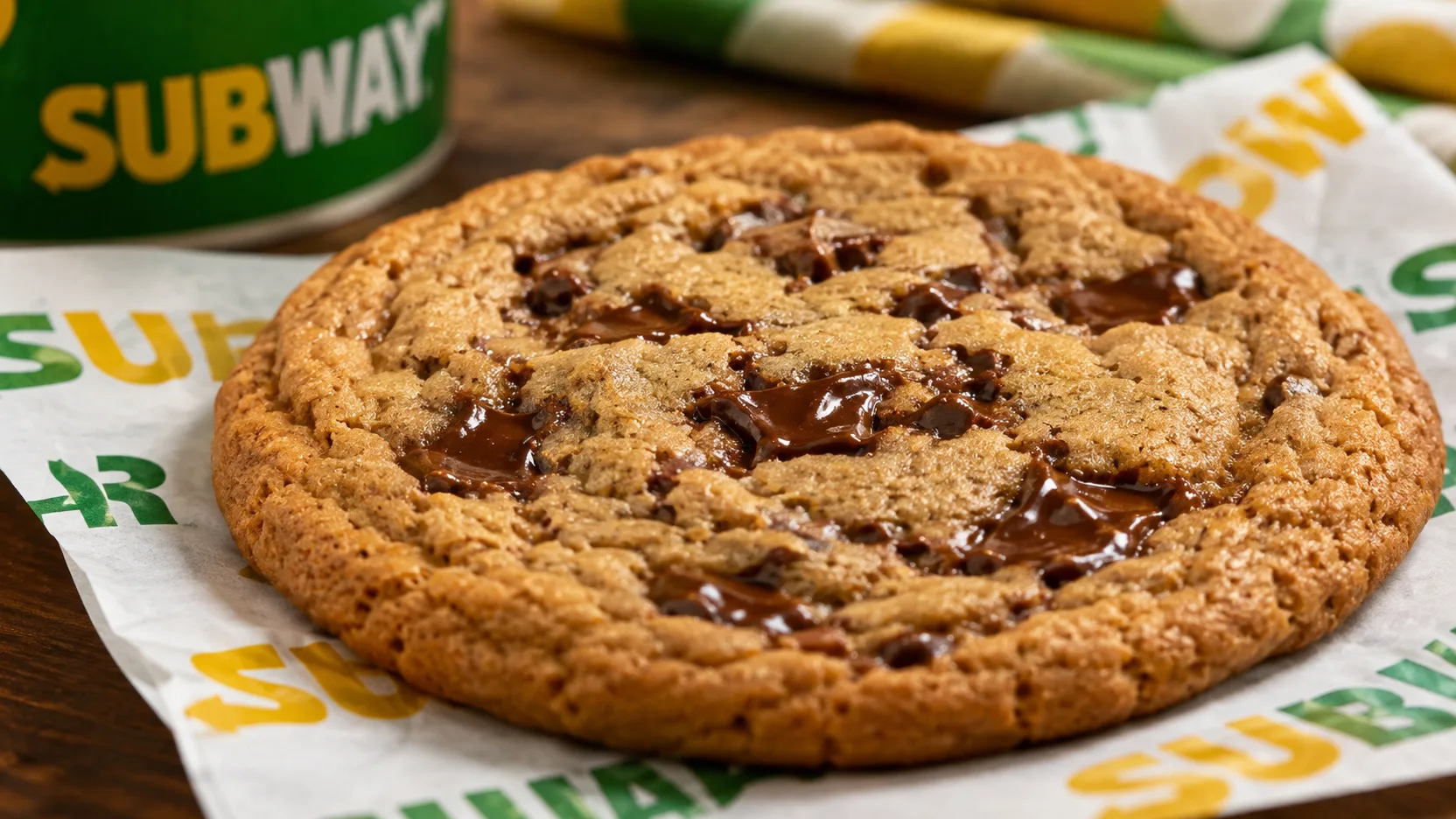 Cookie grande e dourado com pedaços de chocolate derretido, bordas assadas e centro macio, servido sobre papel de lanchonete com embalagem verde ao fundo.