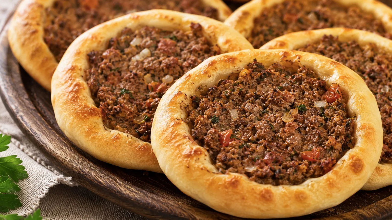 Esfihas abertas de carne assadas sobre tábua de madeira, com bordas douradas e recheio de carne moída, tomate, cebola e ervas.